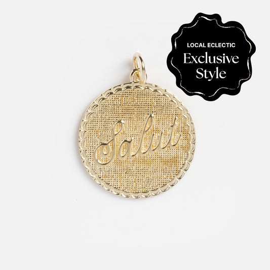 The Zahava x Local Eclectic Globetrotter Charm is a round yellow gold filled pendant with a textured background and Salut in cursive, inspired by travel and available as a Local Eclectic Exclusive Style.
