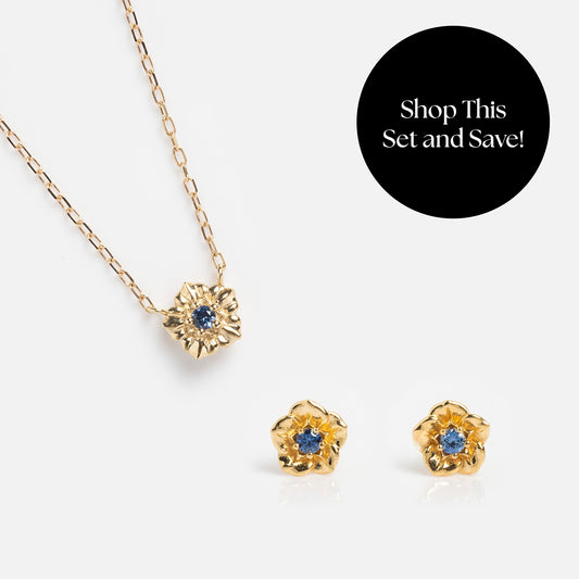 The Solid Gold Birth Flower Gift Set features a flower pendant necklace and matching earrings with blue gemstone centers, displayed on white. A black circle reads, “Shop This Set and Save!”—a perfect gold gift for someone special.