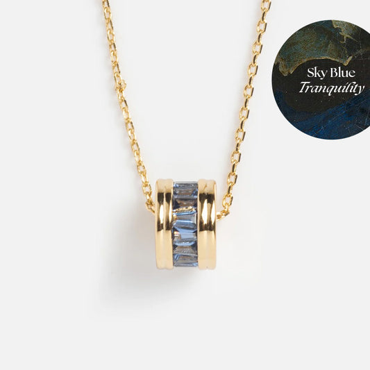 The Color Theory Charm Necklace is a gold-plated necklace with a round pendant featuring rectangular sky blue CZ stones. A circular label in the top right corner reads “Sky Blue Tranquility” on a blue and gold background.