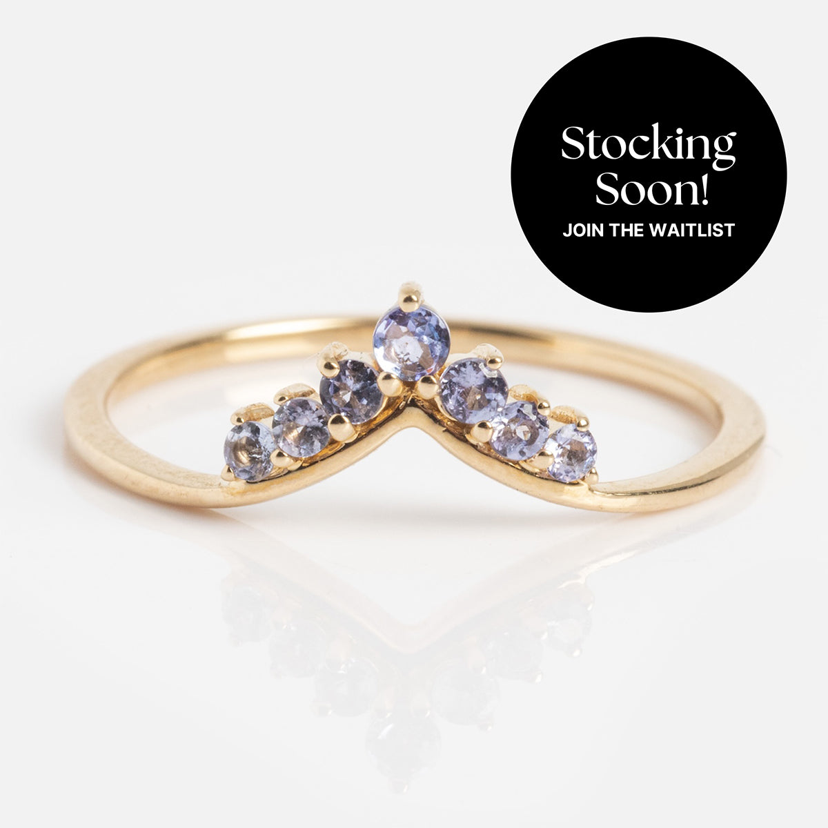 The Solid Gold 2025 December Capsule Delicate Arch Tanzanite Stacking Ring features a chevron design with five 1.5 mm Tanzanite stones. A black circle in the top right reads: Stocking Soon! Join the waitlist.