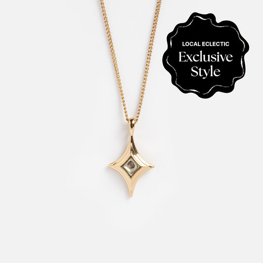The Stellina Pendant Necklace features a diamond-shaped design on a brass base with 14k gold plating and a small central gemstone. Displayed on white, the black label states “Local Eclectic Exclusive Style” in white text.