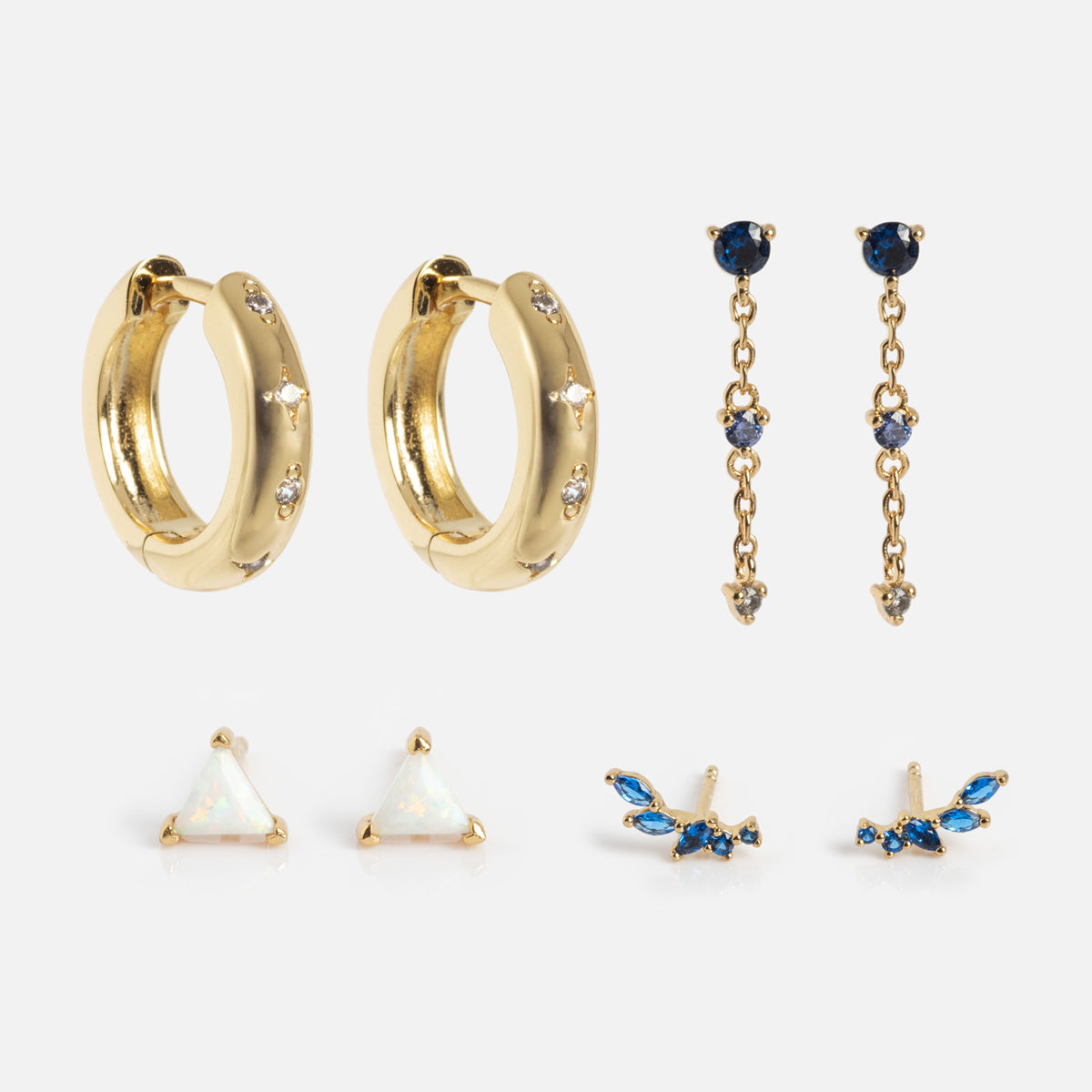 The Bold Birthstone Earring Set includes four pairs: gold hoops with clear stones, dangling gold earrings with blue and clear stones, gold triangle studs with white centers, and small blue/gold leaf-shaped studs.
