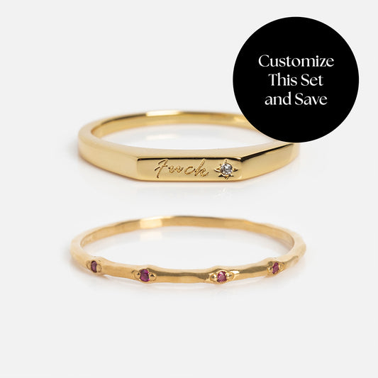 The Solid Gold Tiny Fuck Birthstone Stacking Ring Set features two gold rings: one with a small diamond and engraved text, the other accented with tiny red birthstones. A black circle in the corner reads, Customize This Set and Save.