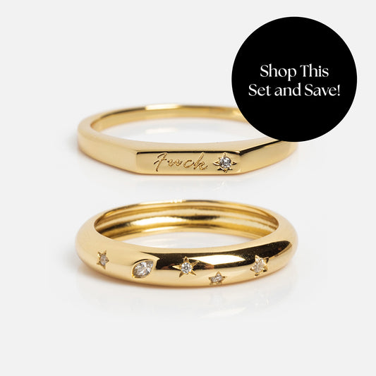 The Tiny Fuck Bold Stacking Ring Set includes two 14k gold-plated rings: one with star engravings and gems, and the other with “Fuck” engraved plus a gem. A black circle reads, “Shop This Set and Save!”.