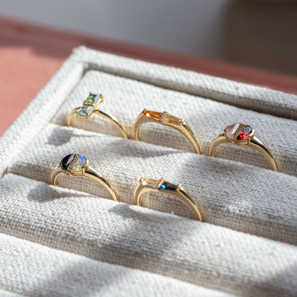 The Solid Gold Toi et Moi North South Ring, featuring colorful gemstones like blue topaz and aquatic agate, shines in a cream-colored fabric jewelry box as sunlight highlights its unique cut and sparkling details.