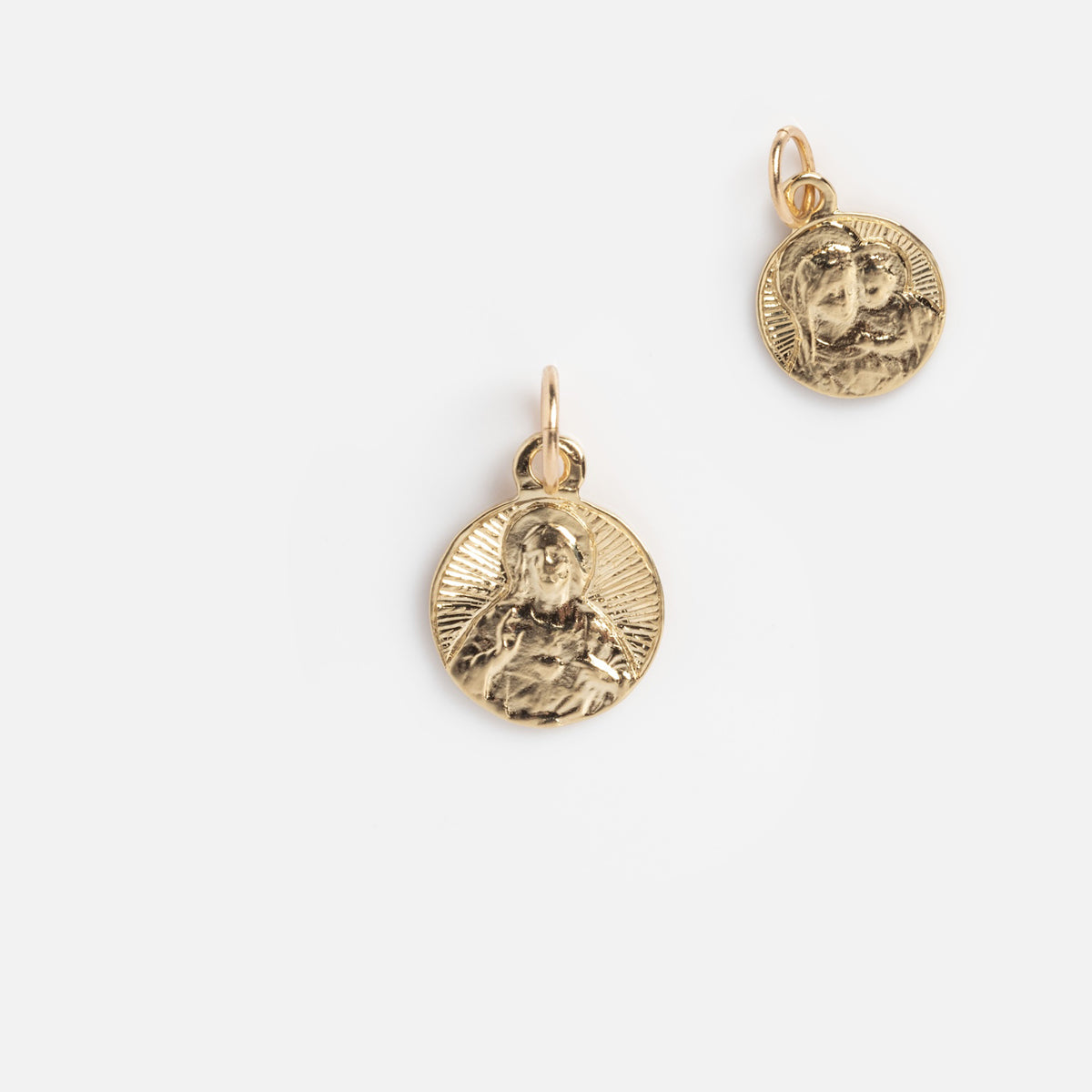 Charms by Charlie and Marcelle: two gold pendants with embossed religious figures and radiant designs, each featuring a small loop for attaching to a necklace or bracelet, displayed on a plain white background.