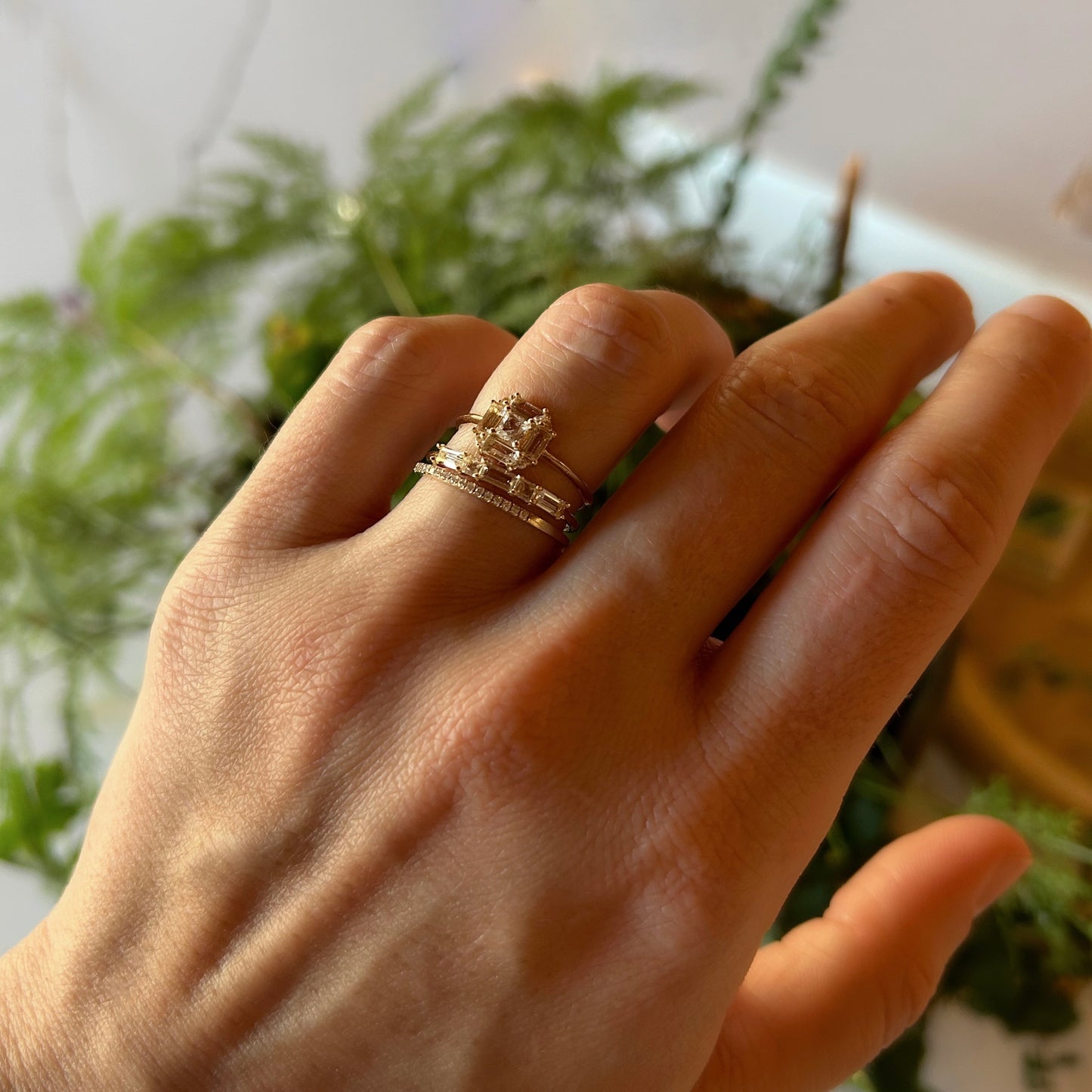 A hand displays three gold rings, including the 14k Vintage Inspired Art Deco Engagement Ring set with diamonds, against a blurred green leafy background.