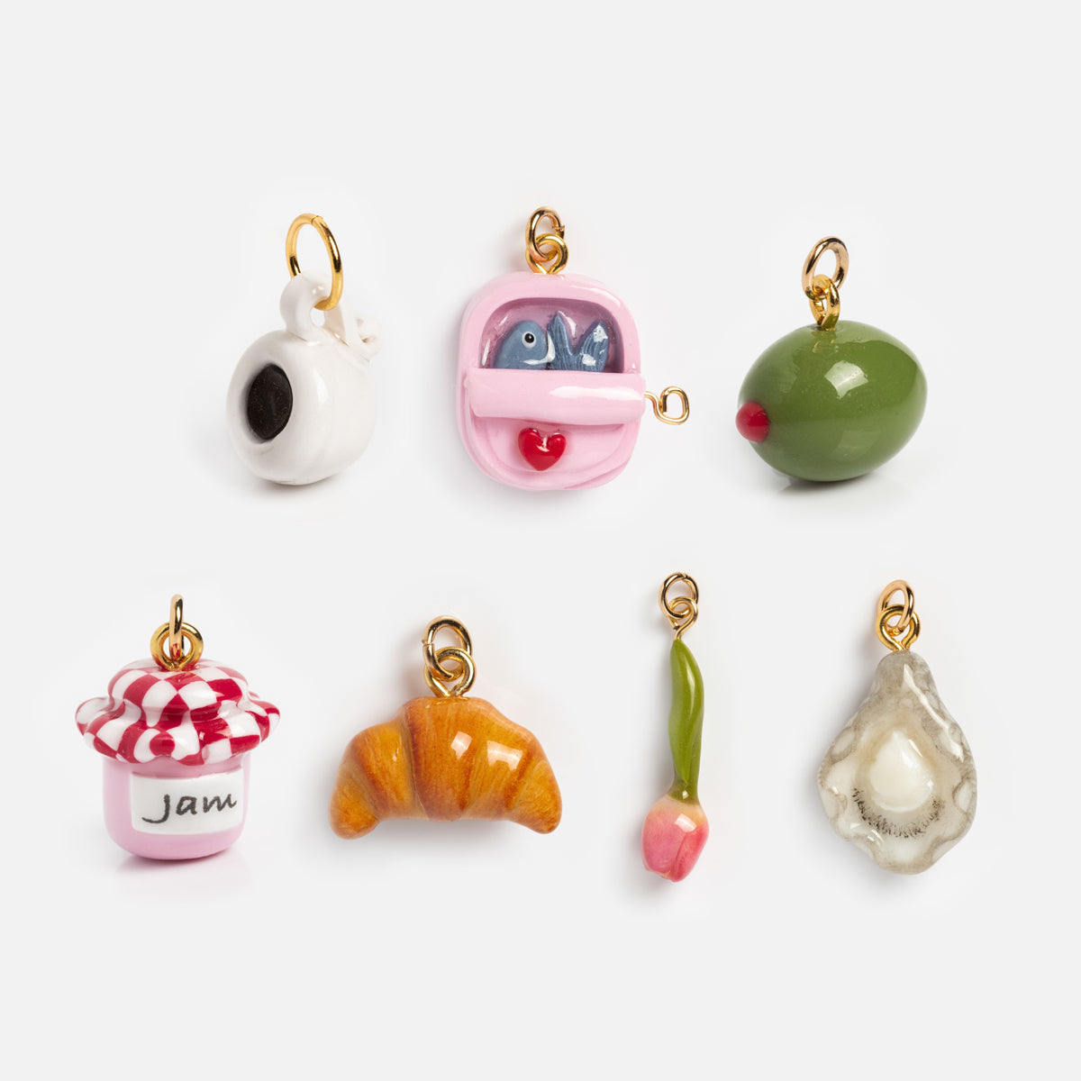 Seven small handmade clay charms from Charms by Warm Clay Co—a teapot, sardines tin, olive, jam jar, croissant, tulip, and oyster—each with a gold loop. Perfect as unique necklace charms for your collection.