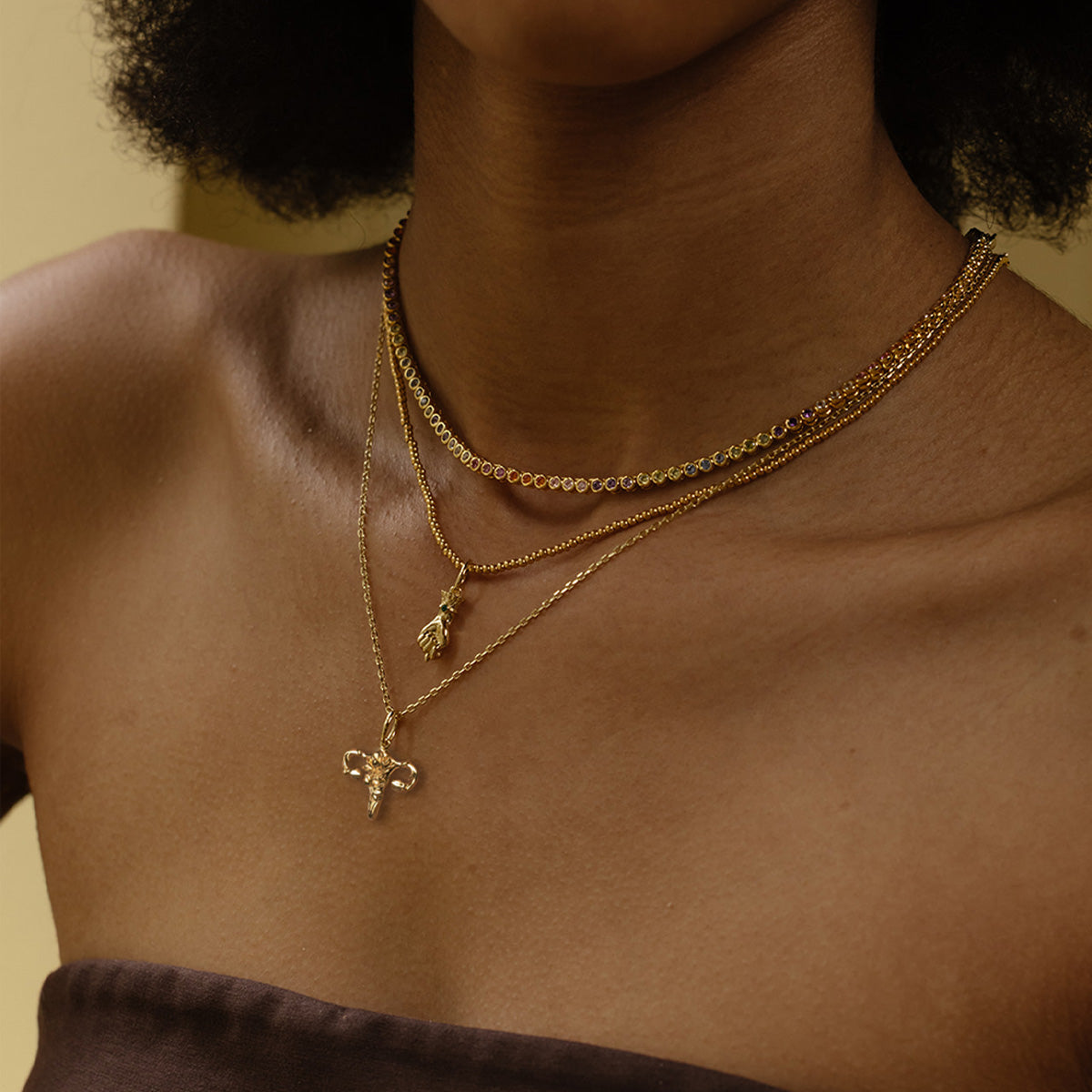 A person in a strapless brown top showcases three 14k yellow gold plated brass layered necklaces with unique CZ pendants against a soft yellow background. Get a Free Gift With Orders Over $200—choose your charm!.