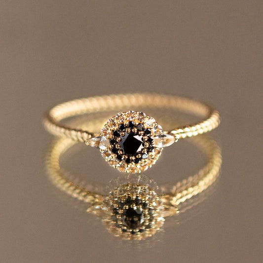 The 9k Black Diamond Halo Ring (Sample Size 7) features a round black diamond at the center, encircled by white sapphires on a textured recycled yellow gold band, beautifully reflected on a smooth surface.