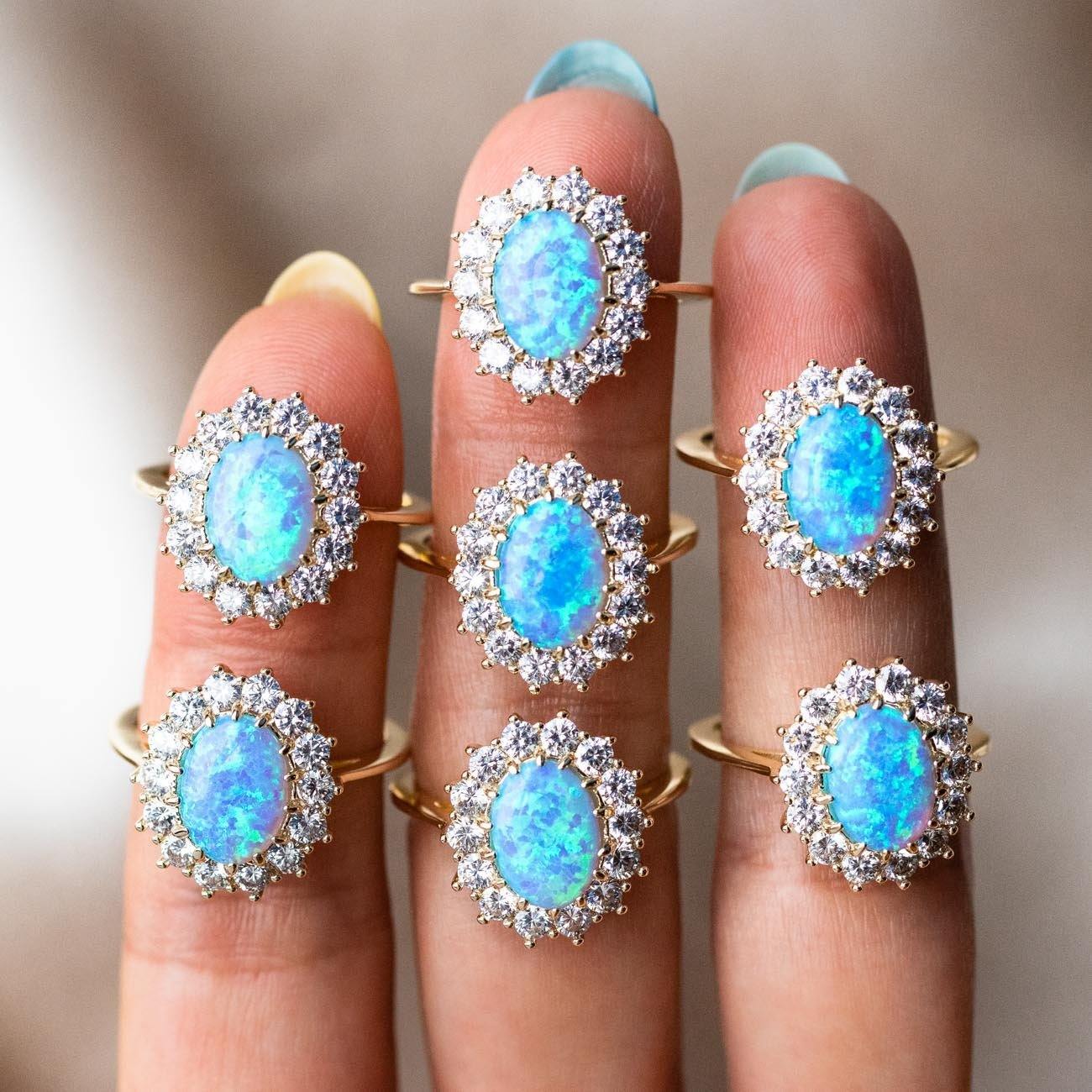 Four fingers with blue-tipped nails display seven Edith Opal Ring samples (size 6), each 18K gold plated with an oval simulated opal and sparkling cubic zirconia in a vintage, ornate design for timeless elegance.