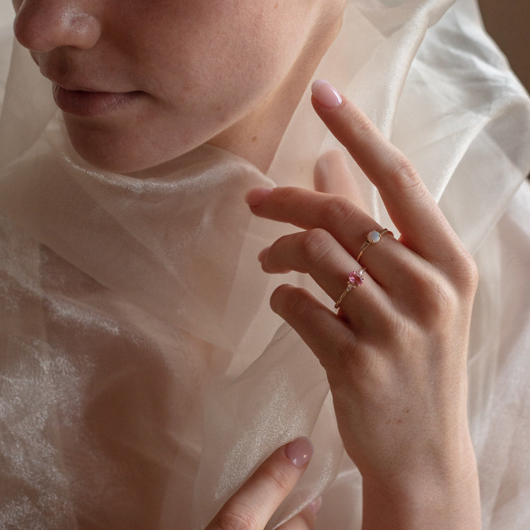 A close-up shows a person in sheer fabric, hands featuring pastel pink nails adorned with delicate 14k Pink Tourmaline and White Sapphire Ring, size 7. The soft lighting highlights the elegant details and gentle style.