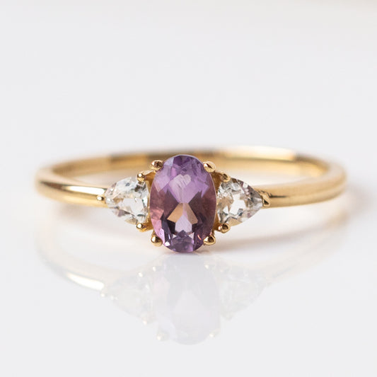 The 14k Amethyst and White Topaz Ring (Sample Size 7) is crafted in recycled yellow gold, with an oval Amethyst center and two pear-shaped white topaz side stones, shown on a white reflective surface.