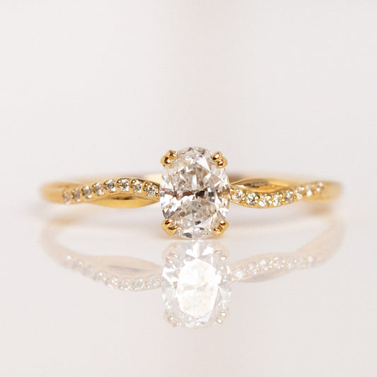 The 9k Oval Cut Diamond Twisted Band Engagement Ring (Sample Size 7) features a lab-created center diamond with small round accents on a twisting gold band, beautifully set against a soft, reflective white background.