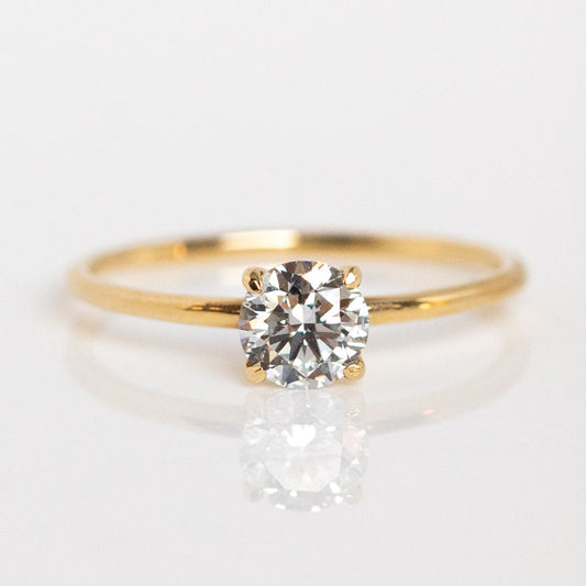 The 9k Classic Round Cut Diamond Solitaire Engagement Ring, sample size 7, features a thin recycled yellow gold band and a single round-cut diamond in four prongs, shown on a reflective white surface.