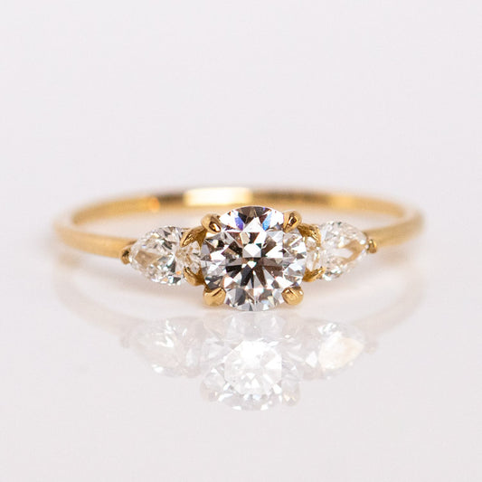 The 9kt Round Cut Diamond Trio Engagement Ring, sample size 7, features a lab-created round diamond with two pear-shaped accents, expertly crafted from recycled solid yellow gold and shown on a reflective white surface.