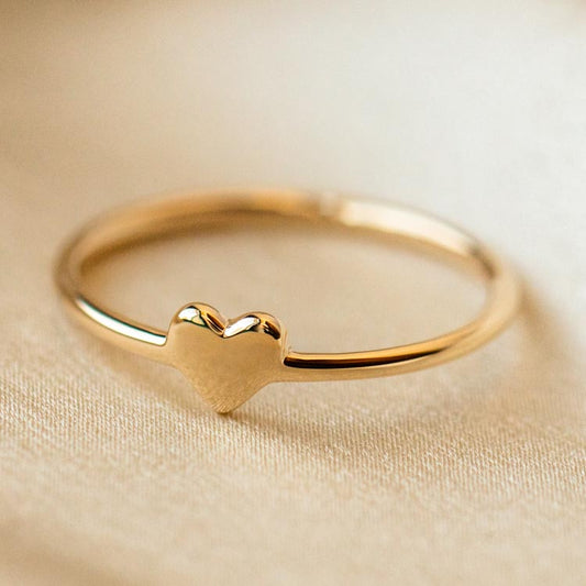 The Solid Gold Engravable Heart Ring Sample Size 9 is a delicate 9k yellow gold ring featuring a small heart-shaped design atop a 1.2 mm band, shown resting on soft beige fabric.