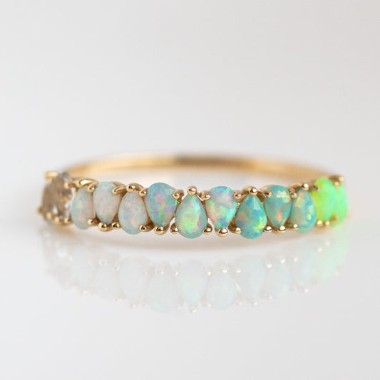 The Solid Gold Ombre Birthstone Ring Opal Sample is a 9k yellow gold ring with a row of pear-shaped simulated opals in ombre shades—white, blue, green, and yellow—set delicately in prongs on a reflective surface.