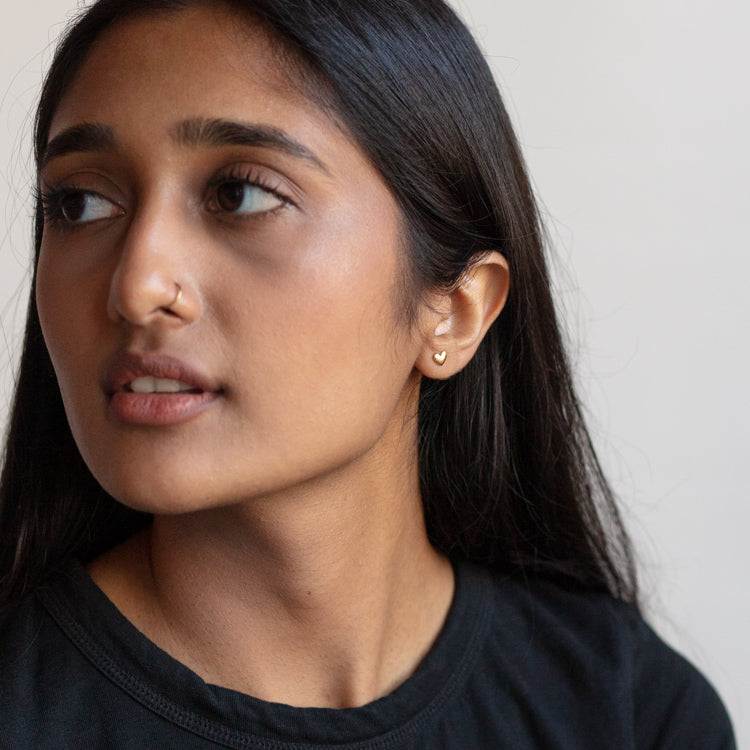 A woman with long dark hair and a nose ring looks left, wearing a black top and Solid Gold Heart Earrings. The background is plain and light.