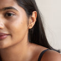 A woman with long dark hair wears a black strap top and displays an elegant ear stack—two delicate gold earrings and the Solid Gold Essential Ear Cuff. She has smooth skin and a calm expression.