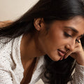A woman with long dark hair and medium skin tone looks down pensively, resting her head on her hand. She wears a white textured blouse and Solid Gold Aquatic Agate Elements Kite Studs in softly warm lighting.