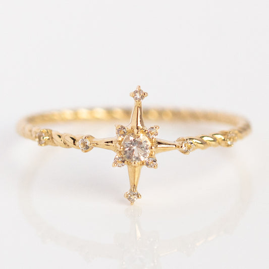 The Solid Gold True North White Sapphire Ring (Size 7 Sample) features a twisted yellow gold band with a central round white sapphire in a star-shaped setting, accented by four small stones at each point.